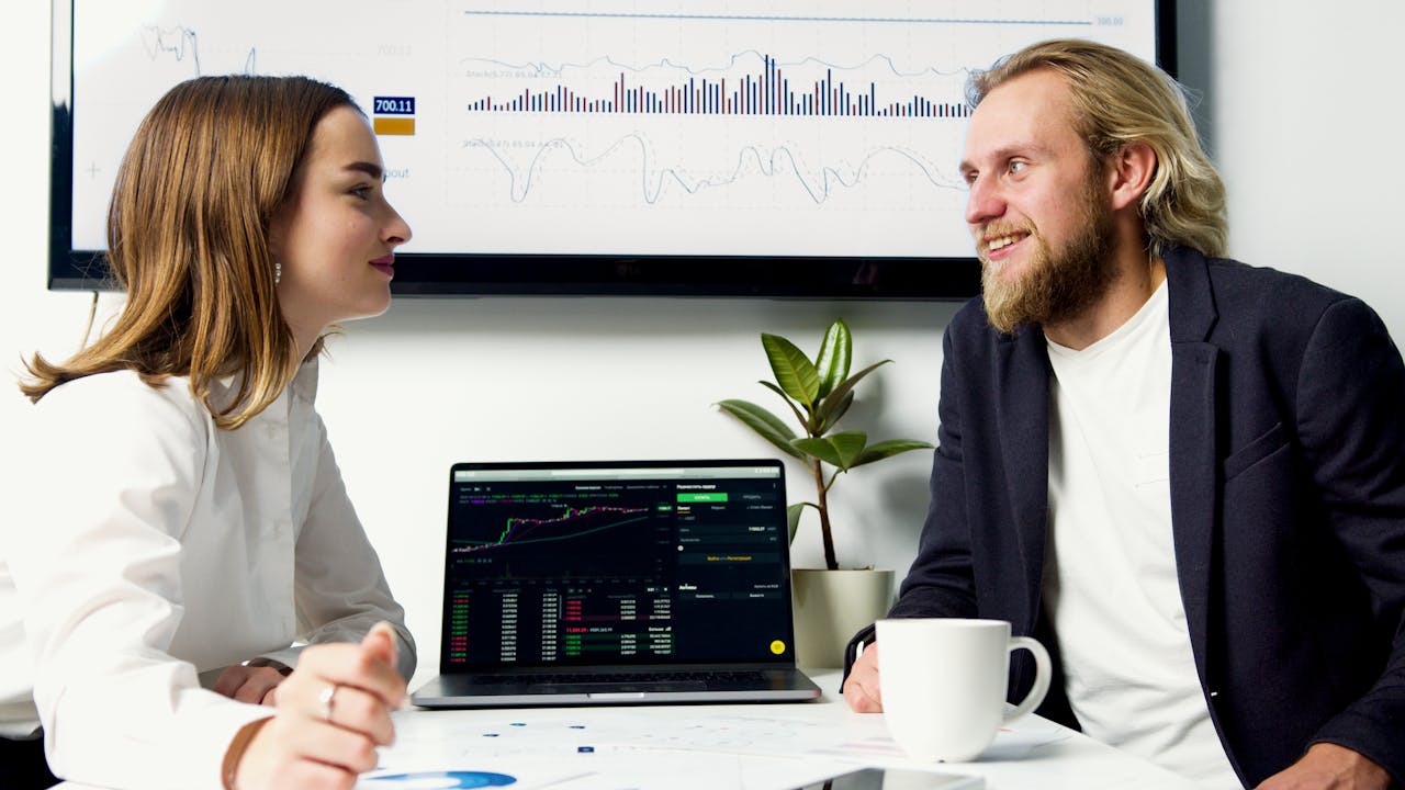 A business meeting with two professionals discussing data on laptops with charts displayed.